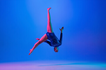 Jumping high. Young flexible girl isolated on blue studio background in neon light. Young female model practicing artistic gymnastics. Exercises for flexibility, balance. Grace in motion, sport