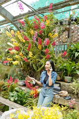 Happy pretty young Asian woman working in greenhouse and talking on phone with customer or contractor