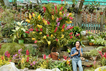 Pretty young Asian woman working in greenhouse and talking on phone when ordering new plants for selling