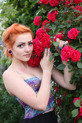 An attractive, romantic, dreamy young girl with red hair on a background of blooming roses. Open air. Summer.