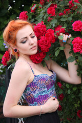 An attractive, romantic, dreamy young girl with red hair on a background of blooming roses. Open air. Summer.