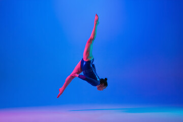 Flying high. Young flexible girl isolated on blue studio background in neon light. Young female model practicing artistic gymnastics. Exercises for flexibility, balance. Grace in motion, sport, action