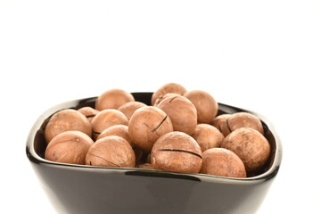 Several whole unpeeled macadamia nuts on a black ceramic plate, close-up, isolated on white.