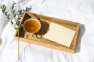 A eucalyptus branch lies on a white sheet beside a tray with a cup of tea and a saucer in a letter in an envelope. The garland is glowing.