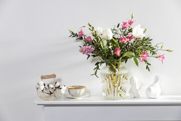 A minimalistic bouquet of white tulips, pink eustoma, hyacinth, eucalyptus in a fluted glass vase on a white panel of an artificial fireplace.