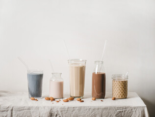 Almond dairy-free milk with blue matcha, berries, coffee, cinnamon, cocoa in bottles and glasses in row on linen tablecloth, white background, copy space. Vegan, healthy, alkaline diet food