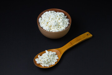 Curd on a black background. Homemade cottage cheese in a deep plate, next to a wooden spoon with...