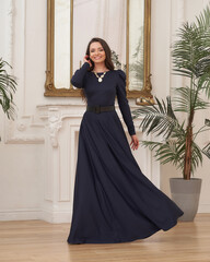 Beautiful pretty woman in long evening dark blue dress posing in luxury white interior. Elegant...