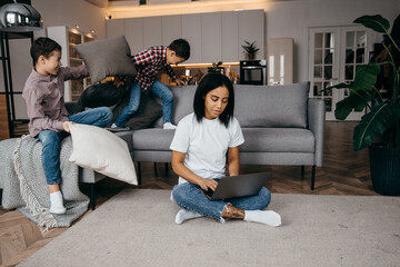 Obraz premium Happy african young mom working at home with laptop and adapting to active noisy sons fighting pillows on sofa
