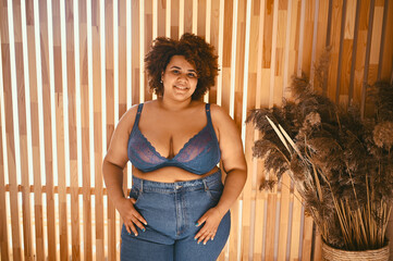 Beautiful stylish curvy oversize African black woman with afro hair posing in bra and jeans on wooden eco friendly background, body imperfection, body acceptance, body positive and diversity concept..