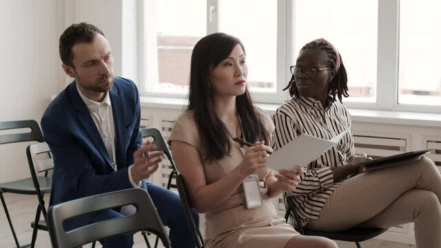 Medium long of Caucasian businessman asking Asian woman sitting on first row of seats, then changing his position to one near her
