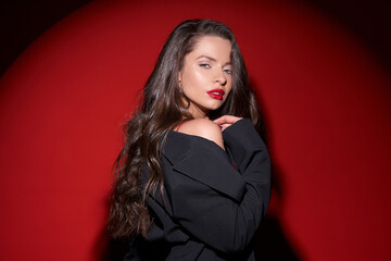 Young beautiful sexy woman with make-up and wavy brunette hair in red silk dress posing against red background in circle of light (spot gobo light)