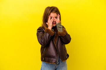 Little caucasian girl isolated on yellow background blink at the camera through fingers, embarrassed covering face.