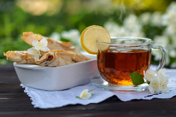 Glass cup with tea and lemon, pastries, croissants, jasmine branches on a wooden table among. Outdoor, picnic, brunch, sunrise, sunset. Selected focus with short DOF. Close up view.