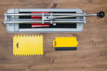a cutter on a wooden surface next to a tile-laying tool, a spatula, and a trowel