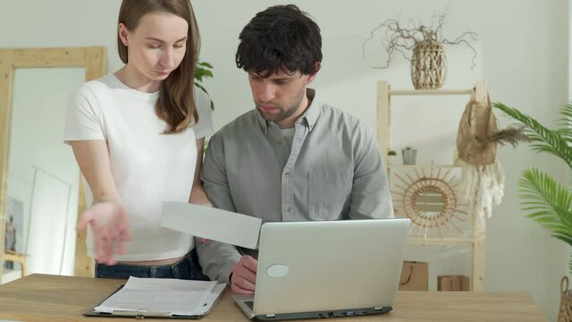 Stressed couple looking frustrated, having no money to pay off their debts, sitting at table with papers, calculator and laptop computer and reading document from bank