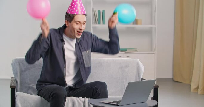 Cheerful active caucasian man at home during lockdown making video call congratulating friend on birthday remotely using video call waving balloons wears festive hat blowing horn whistle creates noise