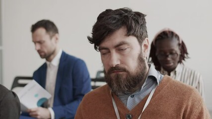 Medium close-up of bearded Caucasian businessman falling asleep on conference, blurred colleagues sitting and chatting on background - Powered by Adobe