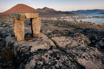 Dusk in a harsh arctic landscape with bare hills and ocean. Inuksuk with Inuit settlement of Qikiqtarjuaq in the background. Broughton Island, Nunavut. © Petr