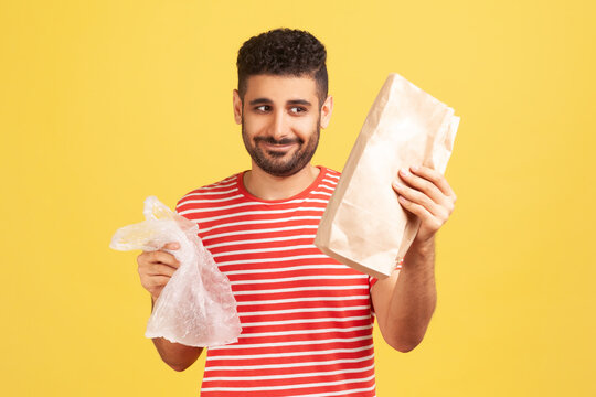 Happy cheerful man with beard in red striped t-shirt holding two bags in hands, looking at paper bag with smile, refusing to use plastic. Indoor studio shot isolated on yellow background