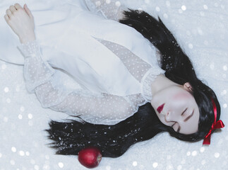Young pretty woman with white skin and red lips is lying on the snow with red poisoned apple.Snow white princess cosplay. © volhavasilevich