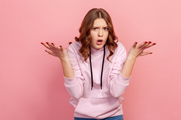Fototapeta premium Portrait of frustrated young brunette female in hoodie spreading hands with shock and misunderstanding, what do you want gesture. Indoor studio shot isolated on pink background