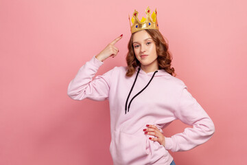 Portrait of ambitious curly haired teenage girl in hoodie pointing finger on crown, feeling...