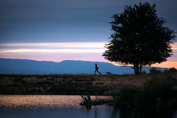 Silhouette of dog and owner running near a lake at sunset.