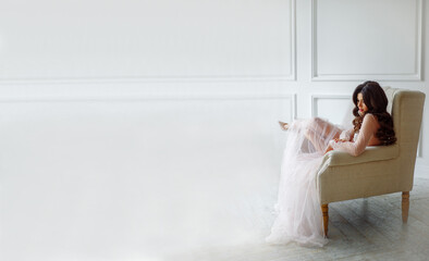 A bride in a white interior in a beautiful boudoir semi-transparent dress. She is sitting on the sofa, she has a beautiful wedding hairstyle and makeup