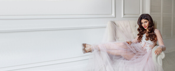 A bride in a white interior in a beautiful boudoir semi-transparent dress. She is sitting on the sofa, she has a beautiful wedding hairstyle and makeup