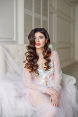 A bride in a white interior in a beautiful boudoir dress. She is sitting on the sofa, she has a beautiful wedding hairstyle