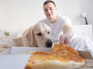 Young man eating pizza at home with laptop and dog. The concept of animals as family members. A guy with a golden retriever is watching a TV show in his bedroom.