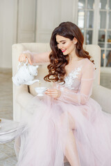 Morning of the bride. She is in a white interior drinking tea. She sits in a chair and pours herself a cup of tea