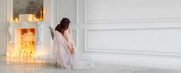 A bride in a white interior in a beautiful boudoir semi-transparent dress. She sits on a chair and hugs her knees. There's a fireplace behind her