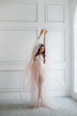 Portrait of a bride in a white interior in a beautiful boudoir semi-transparent dress. She is standing at the window in a beautiful pose
