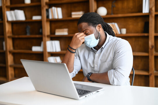 Tired African-American guy wearing mask feel exhausted, a black guy with covered face and eyes closed holds bridge of the nose sitting at the desk in front of a laptop, worrying about his job