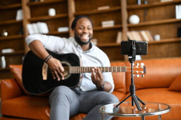 Selective focus at the smartphone on the tripod in front of African guy playing guitar, a vlogger...