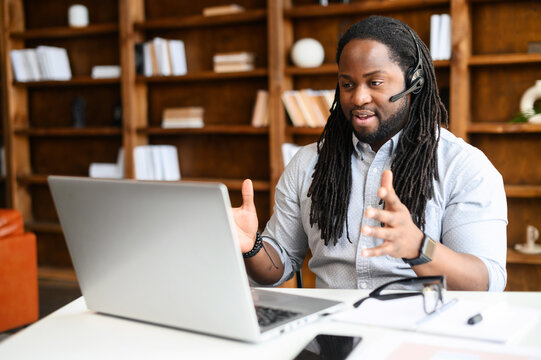Diverse office employees concept. A focused African-American man with a dreadlocks wearing wireless headset is using laptop for online video conversation, talking with customers or coworkers online