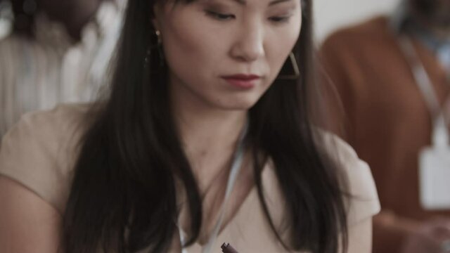 Tracking up medium close-up of young Asian female office worker taking notes in notebook, nodding, looking at invisible speaker, sitting among blurred people