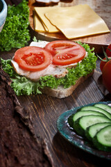 turkey meat sandwich with tomatoes, cheese, green salad and cucumbers on dark wooden table background