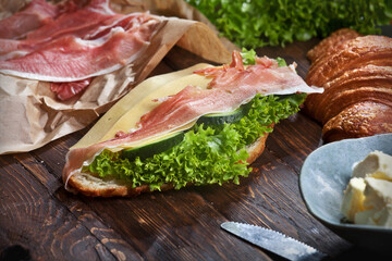 assembling  croissant sandwich with jamon meat slices, green lettuce leaves, fresh cucumbers, cheese and butter