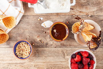 Top view image of a table where hand rolled fresh made waffle cones are dipped into melted chocolate and walnut pieces are sticked on top.  A  bowl of berries and tray of vanilla ice cream is on side