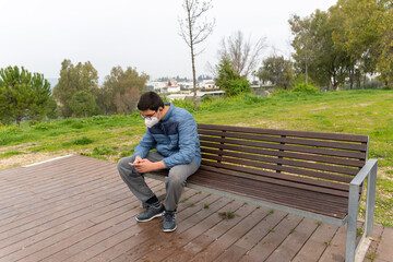 Young boy wearing face mask due to covid19 using his cellphone while sitting in a bench in the park.