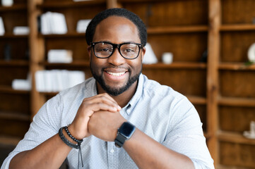 Headshot of friendly and confident African-American guy wearing stylish eyeglasses and smart casual wear, looks at the camera and holds hands in the lock near chin. Employee profile - obrazy, fototapety, plakaty