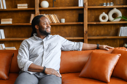 A carefree and cheerful African-American guy rests on the sofa in stylish living room, bookshelves on the background, a smiling multiracial relaxed man with dreadlocks looks away - Powered by Adobe