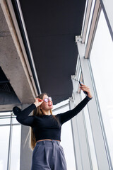 Professional adult businesswoman busy texting on her mobile phone wearing shirt and trousers standing in front of large glass windows.