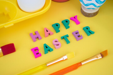 Happy Easter word made of colorful letters with eggs in yellow egg tray, paints and brushes paitning egg in blue waves on yellow background. Creative Easter greeting card.