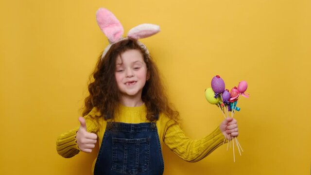 Positive little girl kid wears pink hare ears, points little colored eggs grow, showing thumb up, like gesture, demonstrating approval, isolated over yellow wall. Look at this decorated Easter eggs!