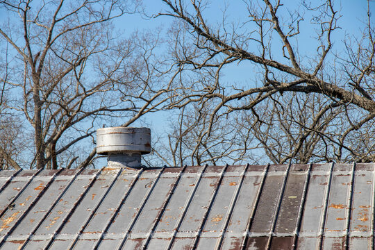 Close-up of a section of vented standing seam metal roof with peeling paint and rust against a background of bare trees and a blue sky. Vintage industrial.