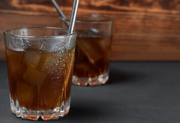 Two low glasses with cold coffee and ice cubes on a black wooden background. Selective focus on the front wall of the glass with highlights and drops. Cocktail iron tubes. Copy space